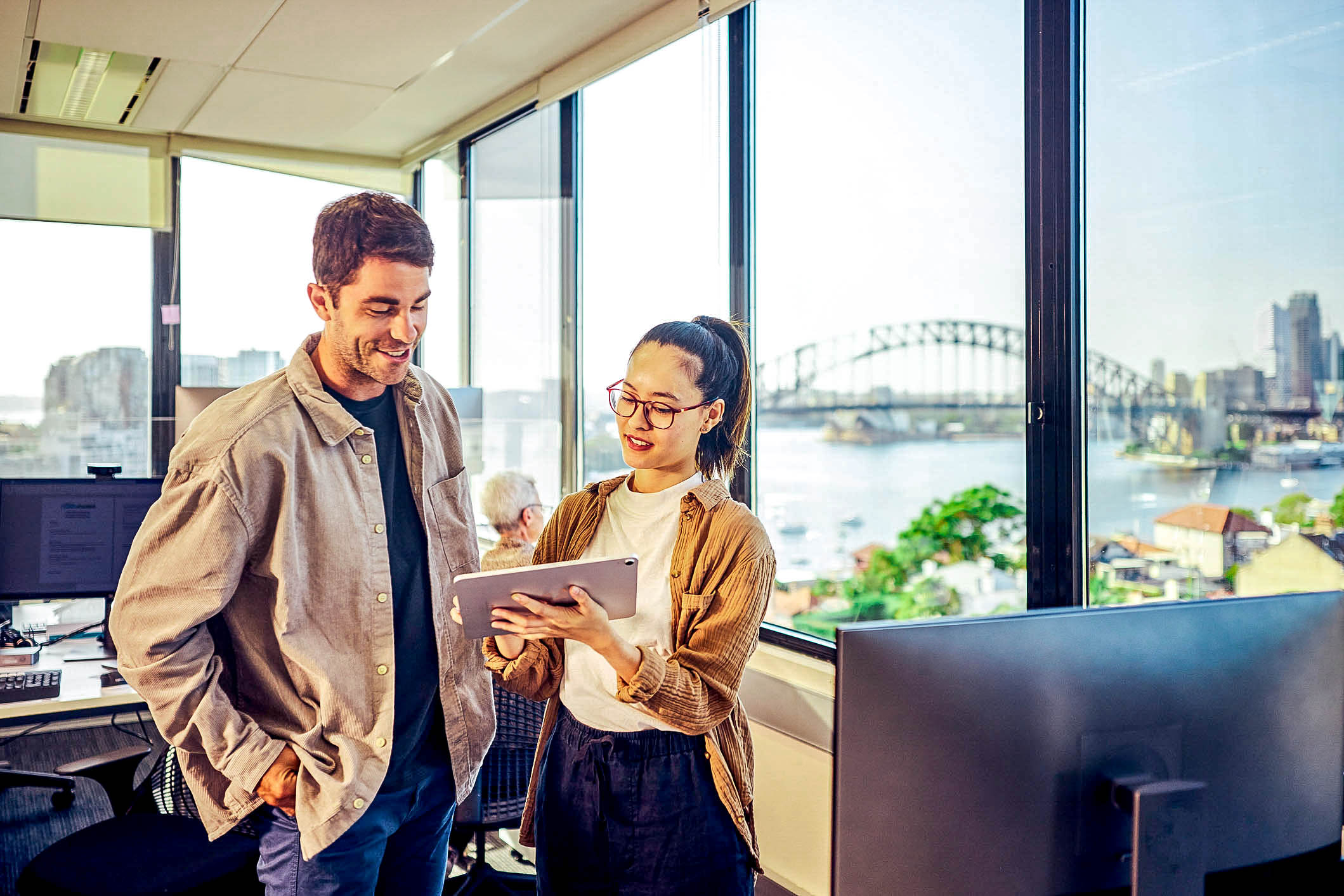 Two employees looking at a tablet, discussing work, with the Sydney Harbour Bridge seen through their office window
